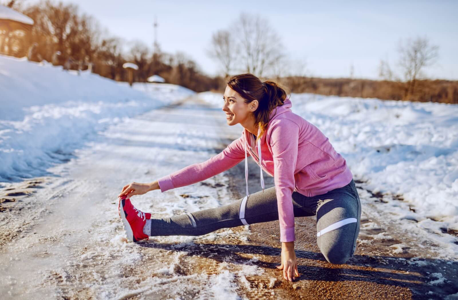 An active young woman stretches outside in the winter time as a break while exercising in the sunshine and snow.