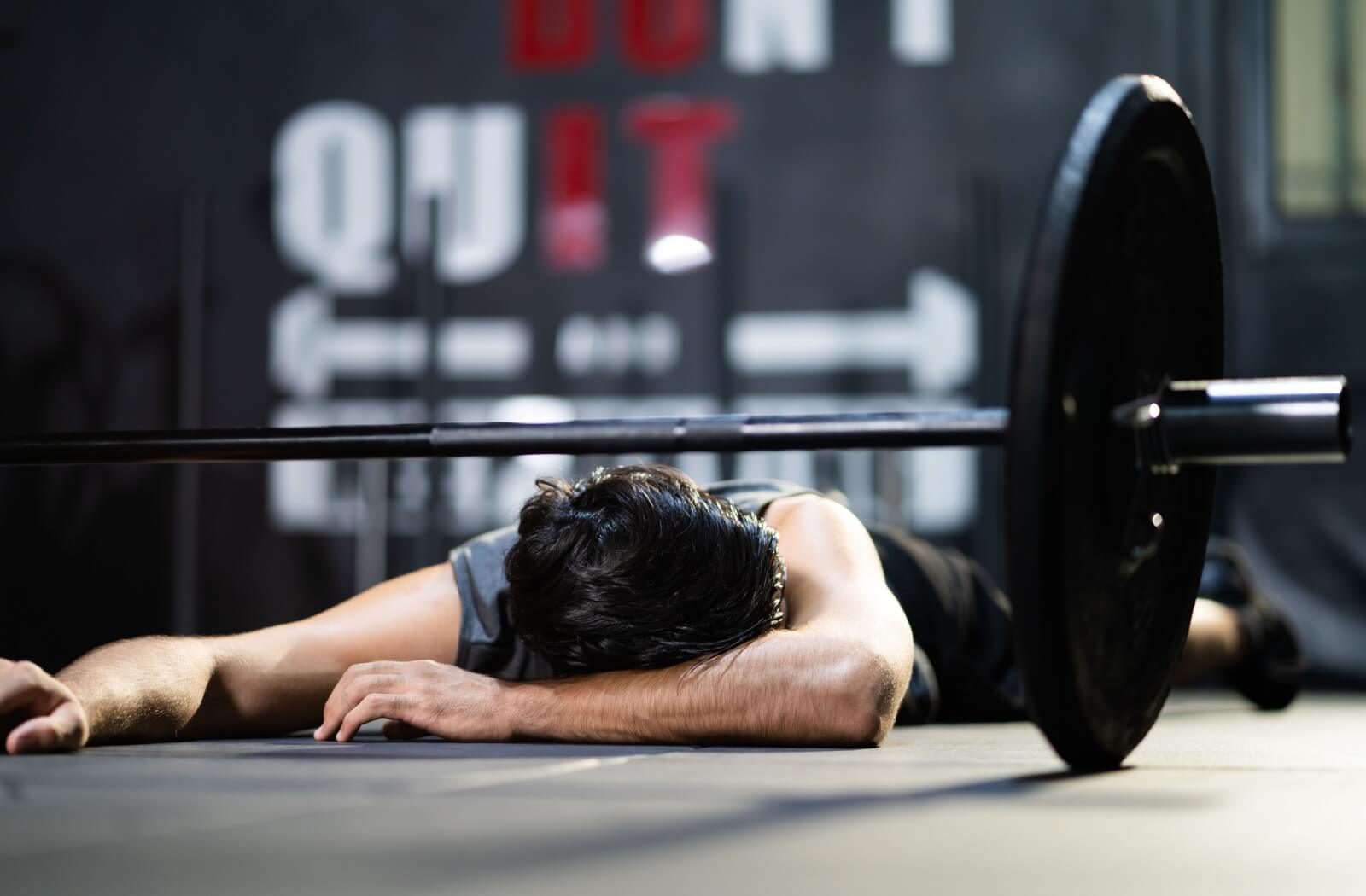 A person lying on the gym floor next to a barbell emphasizing the challenges of fitness consistency