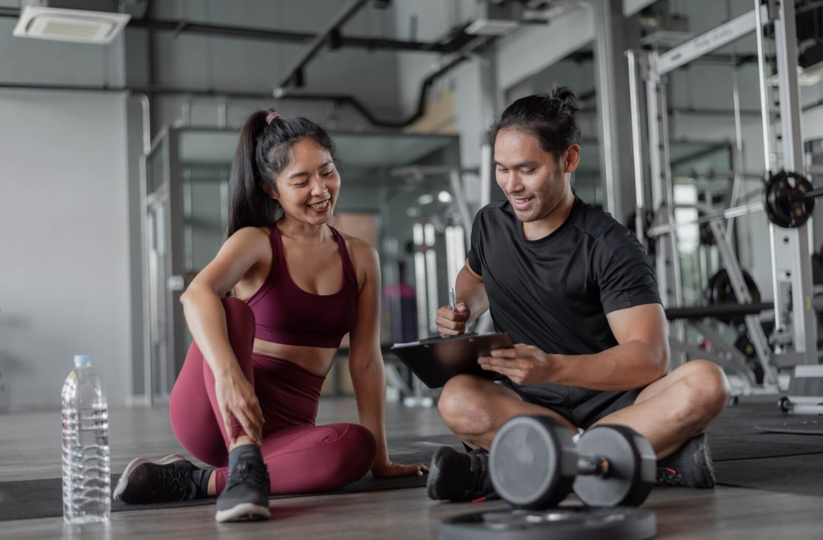 A fitness coach sitting on the gym floor discussing a workout plan with a client promoting exercise consistency and guidance