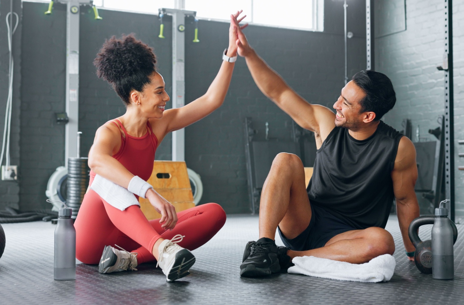 Two people sitting on the ground in a gym high-fiving after finishing a workout.